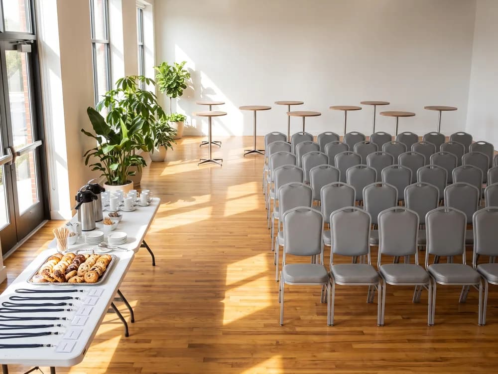Silver banquet chairs in rows in a bright modern event space, wood cocktail tables behind and two 8 ft tables by the door