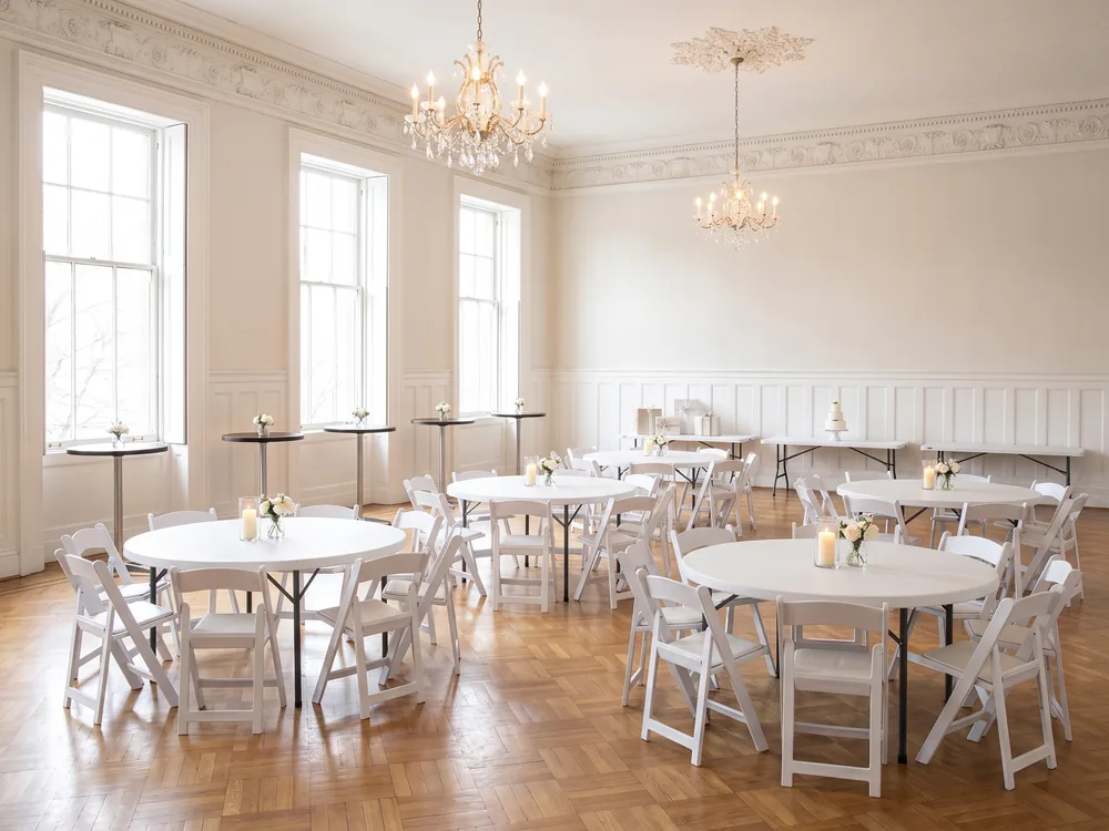 White resin chairs around round tables set for an indoor wedding dinner in an old ballroom, wood cocktail tables by the tall windows