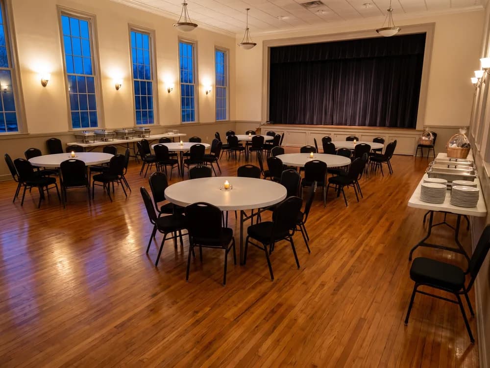 Round tables with black banquet chairs across a banquet hall, two 6 ft service tables along the wall